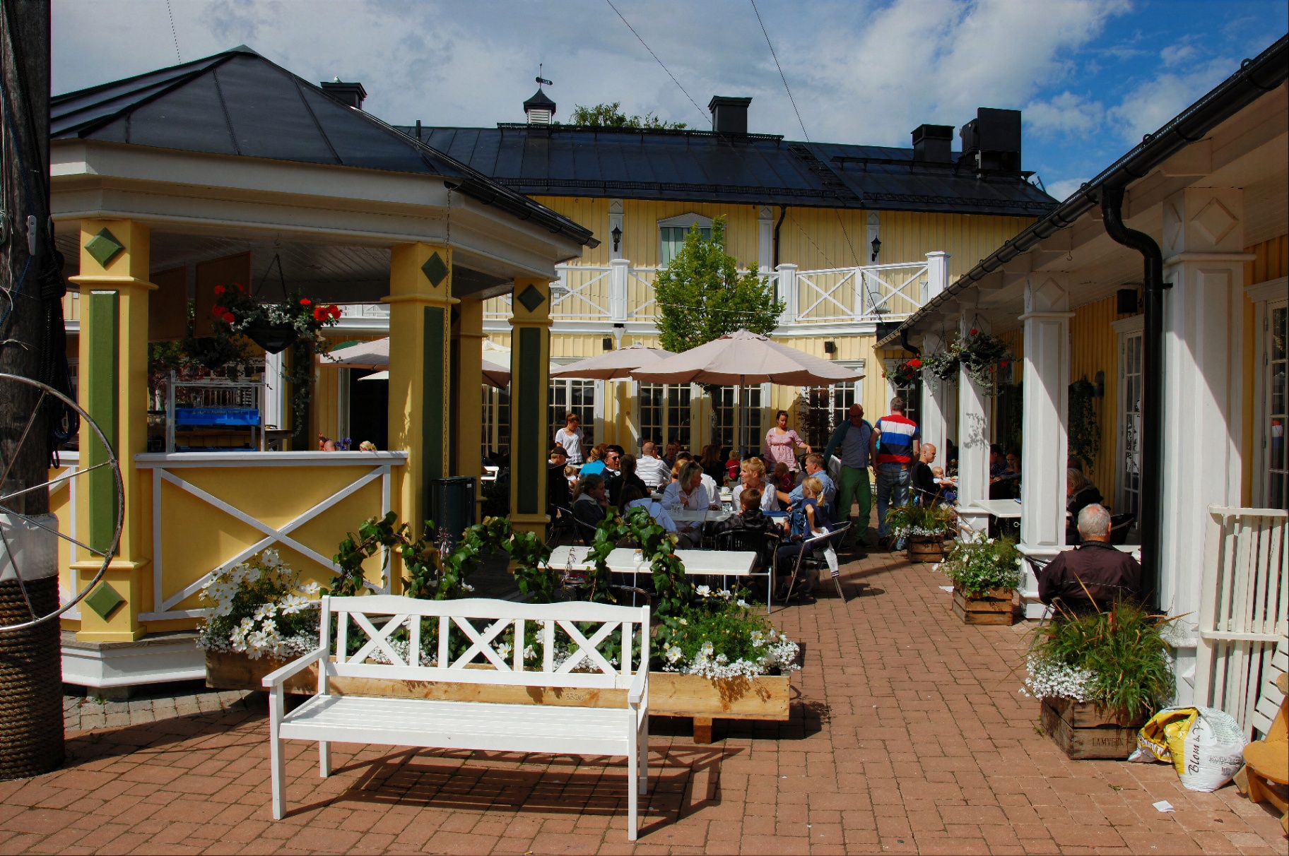 Innergården på Trosa Sjökrog med gäster vid borden, vita parasoll och gula trähus i sommarsol